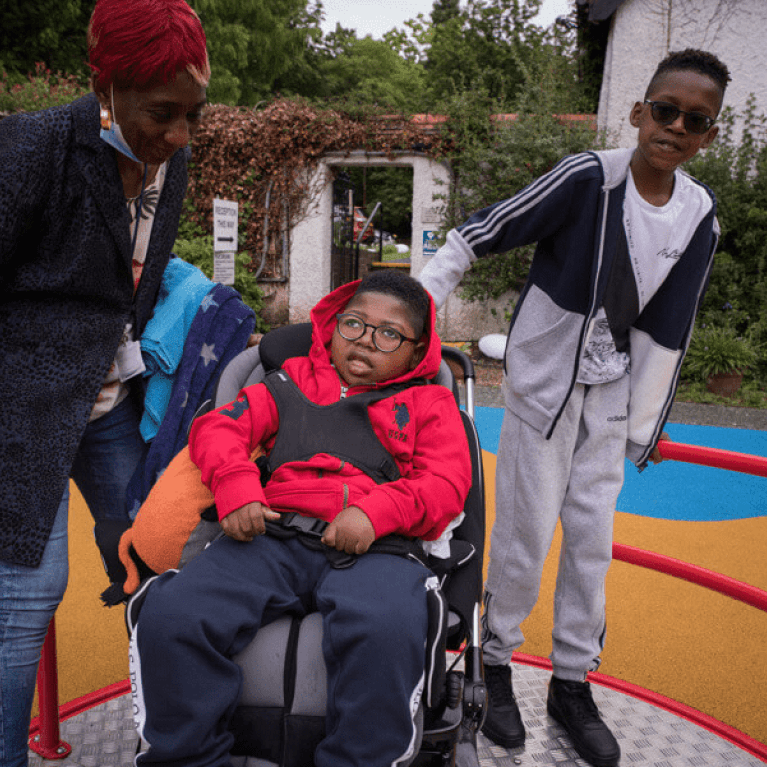 A young boy is on an accessible roundabout in his wheelchair, with his brother and carer